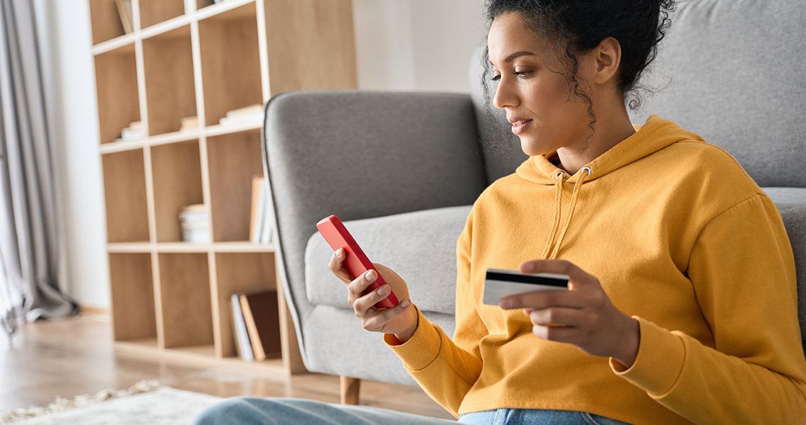 Young adult African American female consumer holding credit card and smartphone sitting on floor at home making payment with app