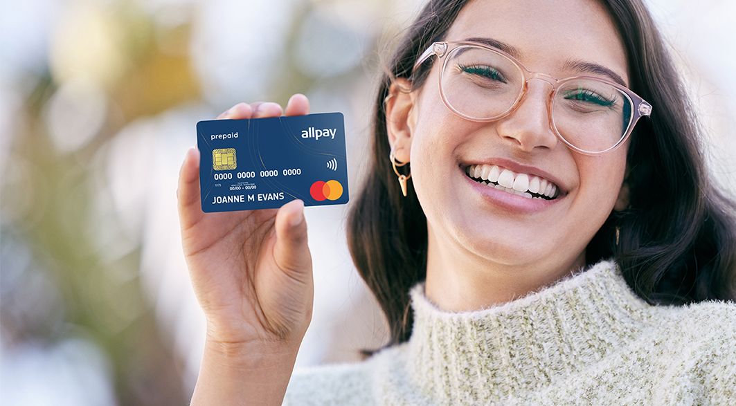 a happy young woman holding up an allpay prepaid card while standing outside.